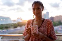Smiling woman holding coffee cup while standing in balcony against sky during sunset — Stock Photo
