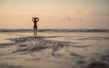 Homem carregando prancha na cabeça enquanto caminhava na praia durante o pôr do sol — Fotografia de Stock