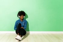 Молодая женщина слушает музыку через наушники, сидя на полу на зеленом фоне — стоковое фото