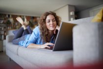 Smiling woman using laptop while lying on sofa at home — Stock Photo