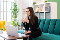Задумчивая молодая женщина держит чашку кофе, сидя с ноутбуком на диване в квартире лофт — стоковое фото