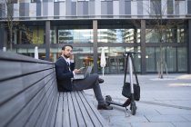 Бізнесмен, який використовує навушники під час сидіння на стіні в місті. — стокове фото
