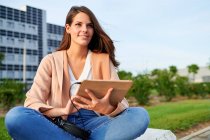 Усміхнена молода жінка дивиться в сторону за допомогою цифрового планшета, що сидить на лавці в громадському парку — стокове фото