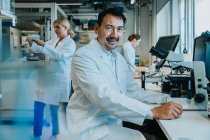 Homme scientifique souriant assis au microscope alors qu'il travaillait en arrière-plan au laboratoire — Photo de stock