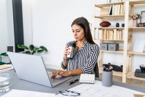 Bella donna d'affari fiduciosa che utilizza il computer portatile mentre beve alla scrivania in ufficio — Foto stock