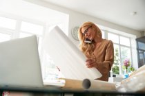 Femme mûre souriante parlant sur le téléphone intelligent tenant carte tout en se tenant à la maison dans la cuisine — Photo de stock