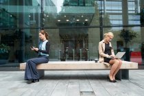Businesswoman and colleague working while sitting with social distance on bench against office building exterior — Stock Photo