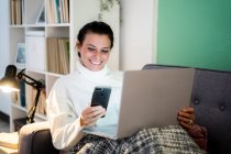 Smiling woman using smart phone and laptop while sitting at home — Stock Photo
