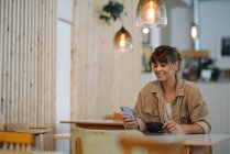 Sonriente mujer propietaria de mensajes de texto en el teléfono inteligente sosteniendo taza de café sentado en la cafetería - foto de stock