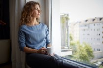 Woman with coffee cup looking through window while sitting on window sill at home — Stock Photo