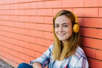 Улыбающаяся женщина слушает музыку через наушники, сидя напротив кирпичной стены — стоковое фото