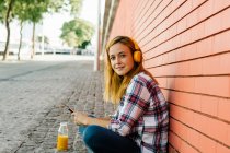 Женщина слушает музыку на смартфоне через наушники, сидя напротив кирпичной стены — стоковое фото