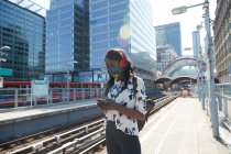 Businesswoman wearing headphones and protective face mask using mobile phone while standing at station platform — Stock Photo