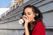 Молодая женщина слушает музыку во время питья кофе из многоразовой чашки в солнечный день — стоковое фото