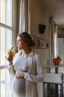 Giovane donna incinta mangiare banana mentre guardando attraverso la finestra a casa — Foto stock