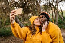 Молода жінка цілує чоловіка, приймаючи селфі через мобільний телефон, стоячи в лісі — стокове фото