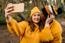 Чоловік цілує жінку, приймаючи селфі через мобільний телефон, стоячи в лісі — стокове фото
