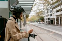 Mittlere erwachsene Frau mit Instrumentenkoffer und Elektro-Schieberoller mit Handy an Straßenbahnhaltestelle in der Stadt — Stockfoto