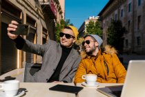 Молодые геи в солнечных очках делают селфи через мобильный телефон, сидя в кафе на тротуаре — стоковое фото