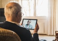 Medico generico maschile che consiglia l'uomo anziano in videochiamata tramite tablet digitale in soggiorno — Foto stock