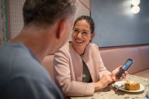 Mujer de negocios sonriendo mientras muestra el teléfono móvil a su colega mientras está sentado en la cafetería moderna - foto de stock
