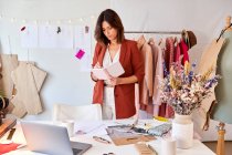 Hermosa mujer diseñadora de moda escribiendo en organizador personal mientras está de pie en el escritorio contra el estante de ropa - foto de stock