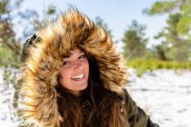 Jeune femme souriante portant un manteau d'hiver à capuche en fourrure fixant debout dans la forêt — Photo de stock