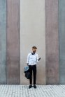 Бизнесмен с портфелем и курткой с помощью мобильного телефона, стоя напротив стены — стоковое фото