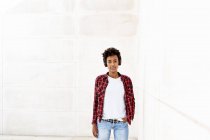 Уверенная молодая афро-женщина слушает музыку, стоя с рукой в кармане у белой стены — стоковое фото