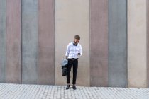 Средний взрослый бизнесмен с помощью мобильного телефона, стоя с портфелем и курткой к стене — стоковое фото