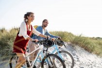 Lächelndes Paar schaut weg, während es mit Fahrrädern am Strand spaziert — Stockfoto
