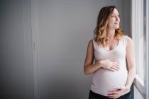 Ragionevole donna incinta con le mani sullo stomaco guardando attraverso la finestra a casa — Foto stock