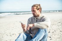 Человек с помощью смартфона, сидя на пляже в солнечный день — стоковое фото
