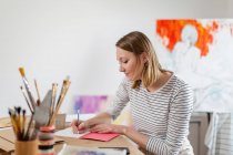 Женщина-художник пишет в книге, сидя за столом в домашней студии — стоковое фото