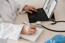 Médico femenino usando tableta digital mientras escribe en el diario en el escritorio en la clínica médica - foto de stock