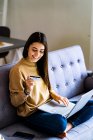 Smiling woman with credit card shopping online while sitting on sofa at home — Stock Photo
