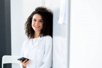 Усміхнена жінка - лікар зі смарт - телефоном, що стоїть на стіні у клініці. — стокове фото