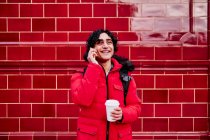 Усміхнений молодий чоловік тримає одноразову чашку кави, розмовляючи по мобільному телефону проти стіни. — стокове фото