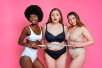Smiling multi-ethnic female models in lingerie measuring waist of worried oversized friend against pink background — Stock Photo