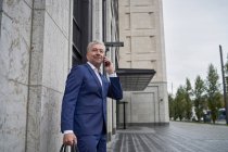 Улыбающийся старший предприниматель разговаривает по мобильному телефону, стоя напротив здания в городе — стоковое фото
