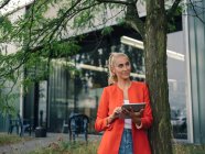 Femme d'affaires avec tablette numérique regardant loin tout en se tenant près de l'arbre à l'extérieur du bâtiment de bureaux — Photo de stock
