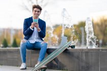 Усмішка молодого бізнесмена з скейтбордом за допомогою смарт-телефону, сидячи на стіні. — стокове фото