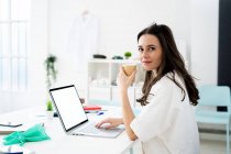 Жінка-лікар випиває каву під час онлайн-консультації за столом у клініці — стокове фото