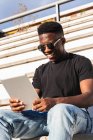 Улыбающийся молодой человек с помощью цифрового планшета, сидя на ступеньках в солнечный день — стоковое фото