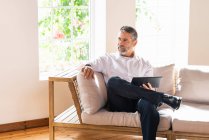 Homme d'affaires réfléchi avec tablette numérique regardant loin tout en étant assis sur le canapé à la maison — Photo de stock