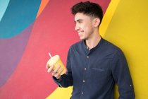 Glücklicher junger Mann hält Einwegbecher in der Hand und blickt gegen bunte Wand — Stockfoto