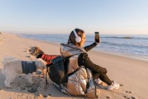 Взрослая женщина делает селфи, сидя с собаками на пляже на закате — стоковое фото