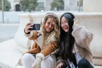Cheerful female friends taking selfie through smart phone while listening music over headphones — Stock Photo