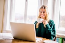 Усміхнена бізнес-жінка сидить з ноутбуком і п'є каву в офісі. — стокове фото