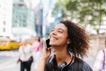 Mulher sorridente com fones de ouvido falando no telefone inteligente na cidade — Fotografia de Stock